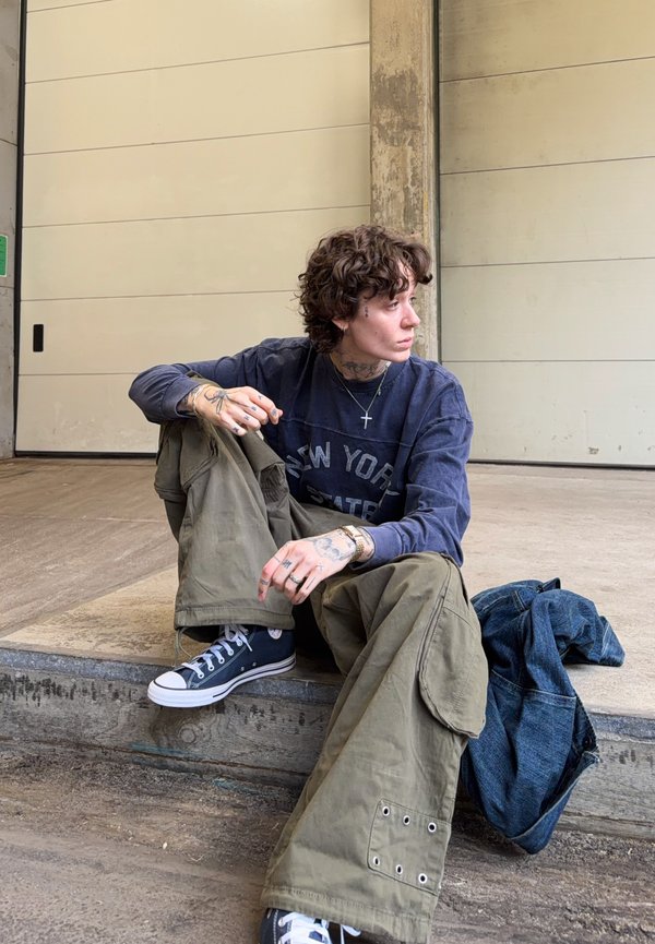 Άτομο με σγουρά μαλλιά και τατουάζ κάθεται σε τσιμεντένιο σκαλοπάτι φορώντας μπλε φούτερ, χακί παντελόνι cargo, μαύρα αθλητικά παπούτσια, με ένα τζιν μπουφάν δίπλα του.