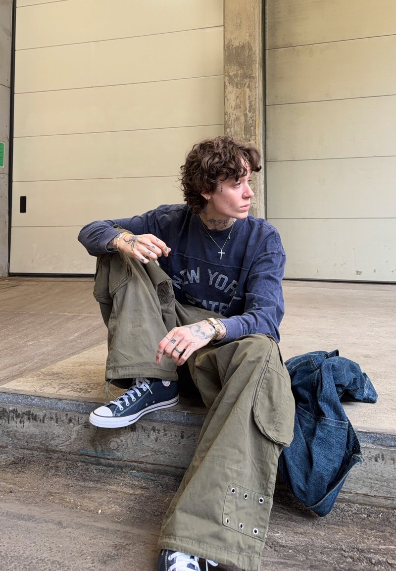 Person med krøllet hår og tatoveringer sidder på betontrin iført navyblå sweatshirt, olivengrønne cargobukser, sorte sneakers med en denimjakke ved siden af dem.