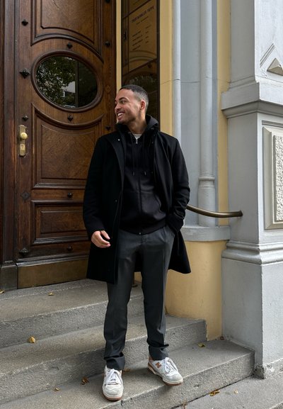 Hombre con un abrigo negro sobre una sudadera negra, pantalones grises y zapatillas. De pie en escalones de piedra junto a una puerta de madera con herrajes de bronce.