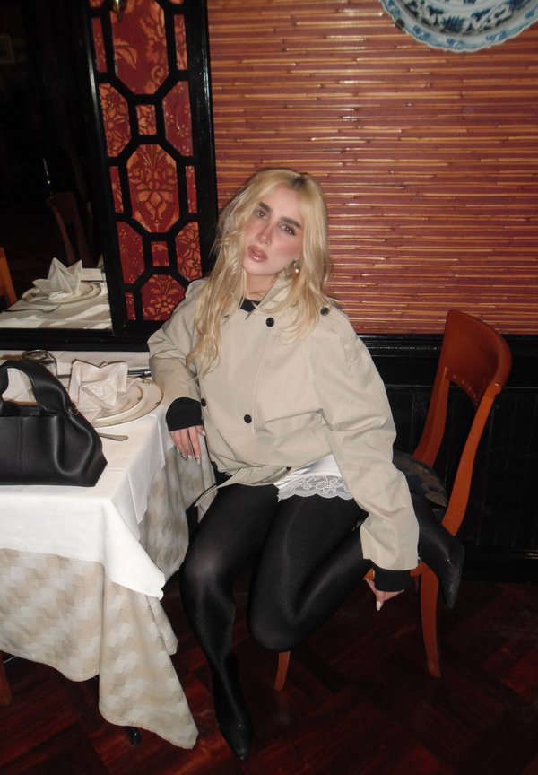Young woman with blonde hair wearing beige jacket and black leggings sits sideways on a chair beside a dining table with plates and a black handbag.
