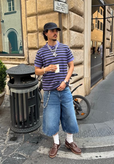 Camisa polo a rayas morada y negra, culottes de mezclilla, calcetines blancos, zapatos marrones. Accesorios incluyen cadenas y una gorra negra. Sosteniendo una taza.