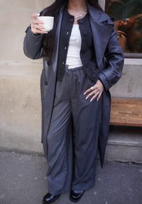 Trench-coat en cuir noir sur un débardeur blanc, associé à un pantalon large à fines rayures grises et des chaussures à plateforme noires. Tenant une tasse blanche.