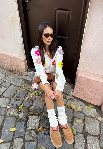 Cárdigan blanco de punto con apliques florales coloridos, zapatillas UGG beige con acentos rojos y calentadores de piernas blancos con canalé. Bolso bandolera marrón.