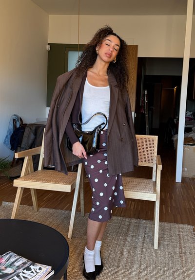 Mujer con blusa blanca, falda de lunares, blazer marrón sobre los hombros, sosteniendo un bolso negro, de pie en una sala de estar con sillas de madera.