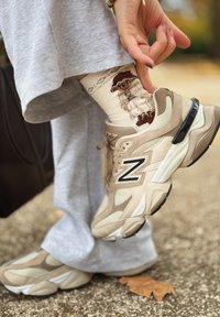 Béžové tenisky New Balance sú vybavené sieťovinou a semišovými detailmi, s čiernym logom. Viditeľné sú vzorované smotanové ponožky s postavičkou.