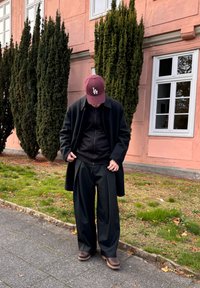 Črni plašč čez črno jakno z zadrgo, široke črne hlače in rjavi čevlji. Bordova kapa z logotipom. Ozadje sestavljajo zelene grmovnice in roza stena.