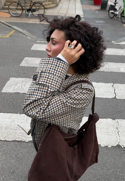 Abrigo a cuadros en tonos marrón y crema con un cuello vuelto, botones negros y un gran bolso de hombro de ante marrón. Anillo de plata visible en la mano.