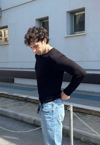 Black long-sleeve ribbed top paired with light blue denim jeans, standing outdoors next to a chain barrier and a stone-paved pathway.