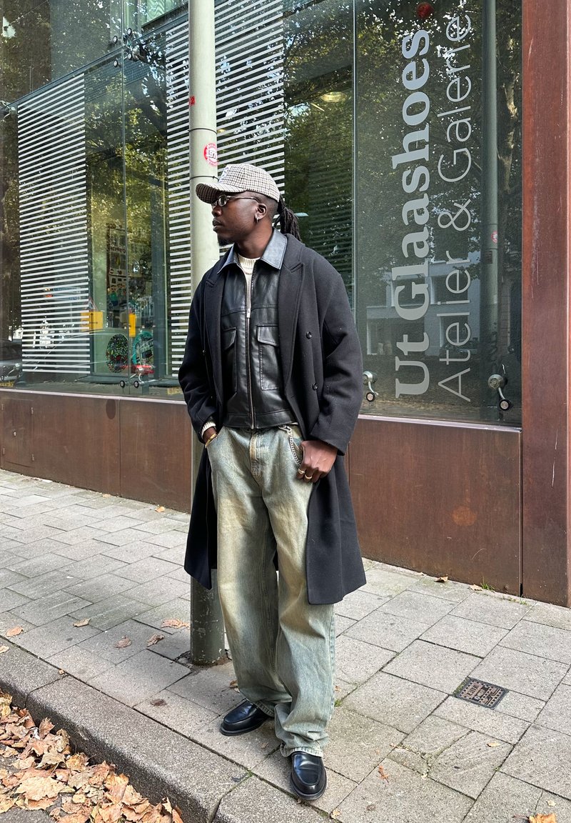 Long black coat over a black leather jacket, teamed with faded blue denim trousers and black shoes. Accessories include a patterned cap and rings.