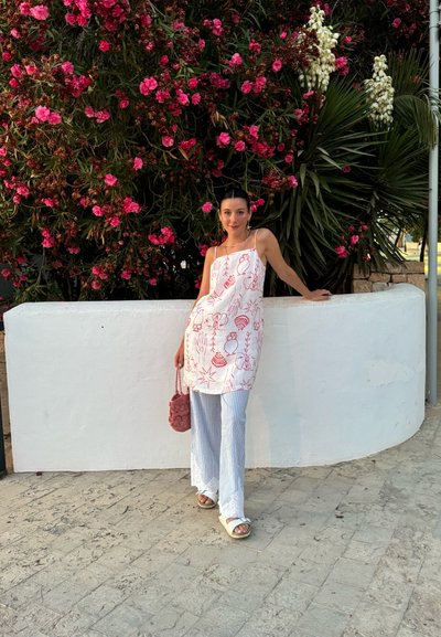 Túnica blanca sin mangas con dibujos rojos, combinada con pantalones de pierna ancha a rayas azul y blanca. Sosteniendo un bolso de mano rosa y esponjoso. Fondo de pared blanca.