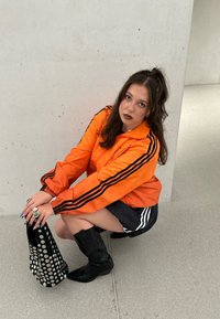 Oranžová tréningová bunda s čiernymi pruhmi, čierne krátke šortky, čierne čižmy až po kolená a perforovaná čierna kabelka so striebornými akcentmi.