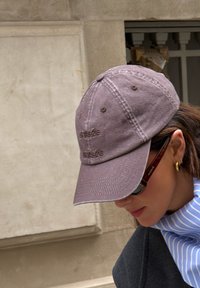 Γυναίκα που φοράει μωβ ξεθωριασμένο καπέλο μπέιζμπολ, γυαλιά ηλίου με μοτίβο χελώνας, χρυσούς σκουλαρίκια κρίκους και μπλε ριγέ κασκόλ, κοιτάζοντας προς τα κάτω.