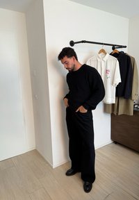 Suéter negro de canalé y pantalones anchos negros, combinados con zapatos negros. El fondo incluye camisas colgadas en blanco, negro y beige.