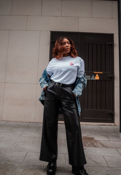 Camiseta azul claro con gráfico rojo, chaqueta de mezclilla, pantalones anchos de cuero negro y botas negras hasta el tobillo, sobre un fondo de pared de piedra neutra.