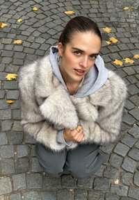 Grå faux fur-kåpe med myk tekstur, stor krage og fremtredende horisontale striper, båret over en grå hettegenser, sammen med grå bukser.