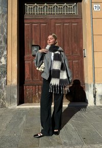 Pelēka vilnas maisījuma pārāk liela jaka, balts tops, melnas platmalu bikses un pelēks kostīms šalle ar franjām. Melni apavi pabeidz izskatu.