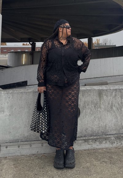 Camisa de encaje negra con botones y falda larga a juego, con un patrón floral. Accesorios incluyen un bolso tote negro con tachuelas y zuecos.