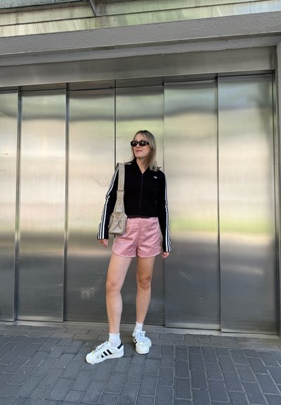Chaqueta negra con cremallera y rayas blancas, pantalones cortos rosas y zapatillas blancas con rayas negras. Bolsa gris, gafas de sol y fondo de pavimento en baldosas.