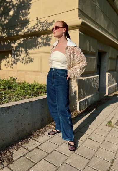 Top blanco sin mangas, cobertura de encaje, jeans de mezclilla azul de tiro alto y sandalias de malla negras. Los accesorios incluyen gafas de sol y pendientes.