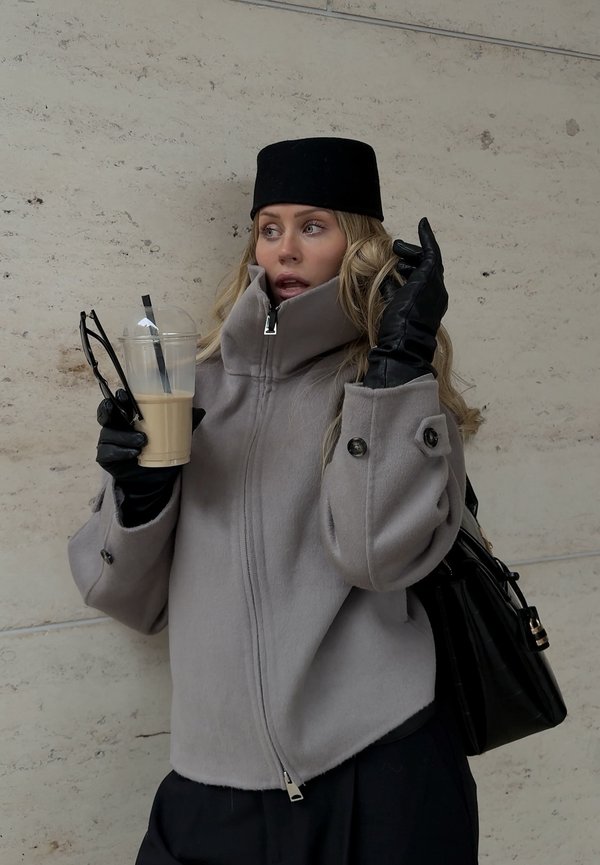 Kvinde med sort hat og handsker iført grå lynlåstrøje, der holder iskaffe og solbriller, stående foran en lys stenvæg.