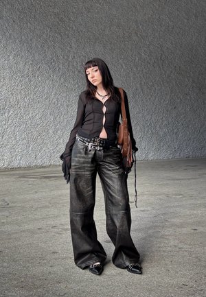 Camisa larga negra transparente de manga larga con abertura en la parte delantera, combinada con pantalones holgados de cuero negro. Los accesorios incluyen un cinturón con tachuelas y un bolso marrón.