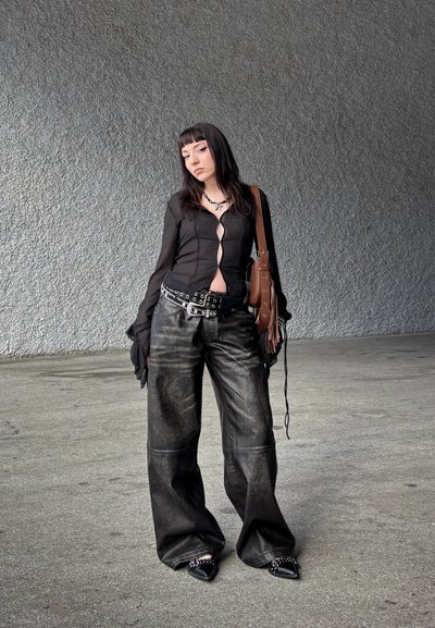 Camisa larga negra transparente de manga larga con abertura en la parte delantera, combinada con pantalones holgados de cuero negro. Los accesorios incluyen un cinturón con tachuelas y un bolso marrón.