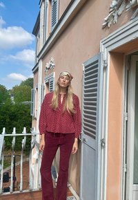 Fließende Bluse mit Blumenmuster in Dunkelrot, kombiniert mit passenden burgunderfarbenen Schlaghosen. Kopftuch mit goldenen Akzenten. Hintergrund besteht aus einer pastellfarbenen Wand.