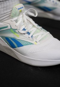 Sapata atlética branca com design em rede, acentos azuis e verdes, superfície texturizada e atacadores brancos, apresentando um logótipo Reebok em destaque.