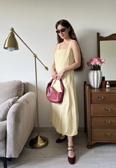 Vestido amarillo sin mangas con escote cuadrado, bolso texturizado rojo y zapatos burdeos. Lampara de pie dorada y cómoda de madera de fondo.