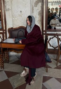 Vinrød ullfrakk over en grå hettegenser, sittende ved et trebord med en svart håndveske. Brune sko med dusker og et skjerf med frynser.