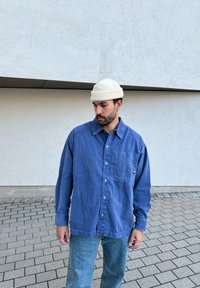 Camisa de algodão azul com botão, com um bolso no peito, combinada com jeans azul-claro e um gorro de malha creme, contra um fundo cinza texturizado.