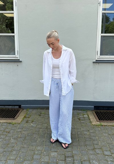 Camisa blanca de botón sobre una camiseta de tirantes blanca, combinada con pantalones de pierna ancha a rayas azul claro y chanclas negras. Fondo gris.