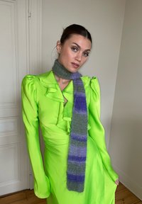 Vestido verde-lima com decote franzido e mangas longas, combinado com uma cachecol cinza fofinho com riscas roxas. Chão de madeira e paredes brancas.