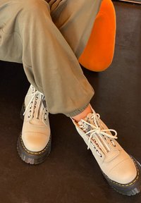 Botas de cuero gris claro con suela negra gruesa, cremallera frontal y cordones blancos; acentuadas con costuras amarillas y detalles texturizados.