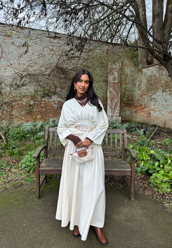 Mujer con cabello largo y oscuro de pie en un jardín, vestida con un vestido blanco sobre una camiseta marrón, sosteniendo un bolso pequeño beige y marrón, con un banco de madera detrás de ella.