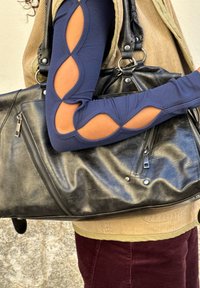 Kék, hosszú ujjú, kivágásokkal ellátott felsőt viselő személy, bézs mellénnyel és piros nadrággal, aki egy nagy fekete bőr kézitáskát hord a vállán.