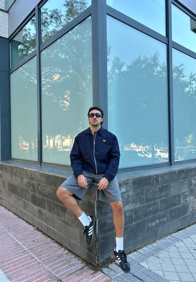 Chaqueta de cierre navy con bolsillos frontales, pantalones cortos grises y zapatillas negras con rayas blancas, sentado en un muro de piedra cerca de grandes ventanales de vidrio.