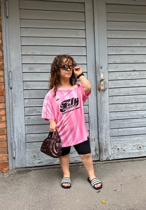 Camisa oversized a rayas rosas con logo negro, pantalones cortos negros, sandalias de diapositivas en blanco y negro, y un pequeño bolso acolchado. Cabello ondulado y gafas de sol.