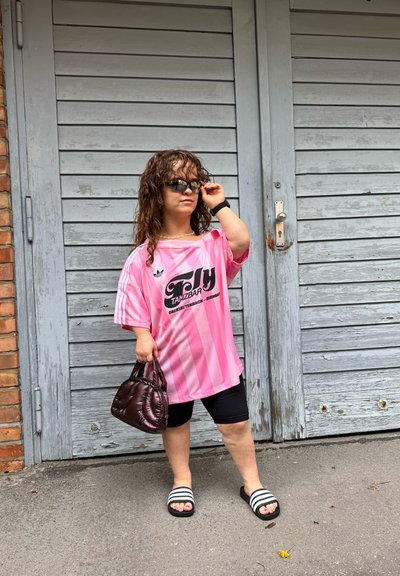 Camisa oversized a rayas rosas con logo negro, pantalones cortos negros, sandalias de diapositivas en blanco y negro, y un pequeño bolso acolchado. Cabello ondulado y gafas de sol.