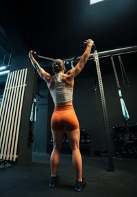 Femme athlétique en débardeur blanc et shorts oranges effectue un exercice avec des bandes de résistance dans une salle de sport aux murs noirs et avec des poids.