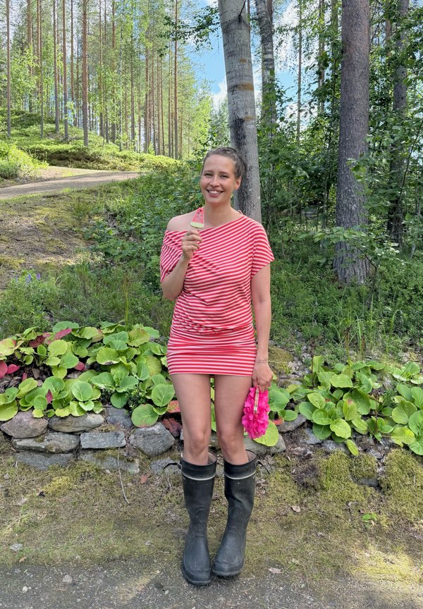 Femme souriante en robe à rayures rouges et bottes noires se tient dans une forêt tenant une glace à la pastèque et un collier de fleurs rose.