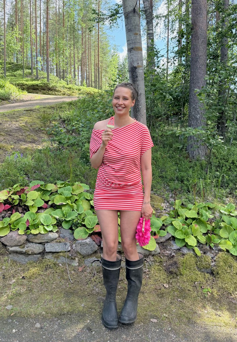 Femme souriante en robe rayée rouge et bottes noires, debout dans une forêt, tenant une glace à la pastèque et un collier de fleurs rose.