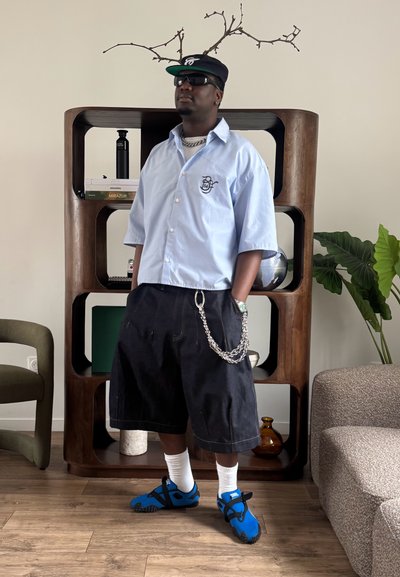 Hombre con camisa azul clara, gorra negra, gafas de sol, pantalones cortos negros largos con cadena, calcetines blancos y sandalias deportivas azules, de pie dentro de una habitación junto a una estantería de madera.