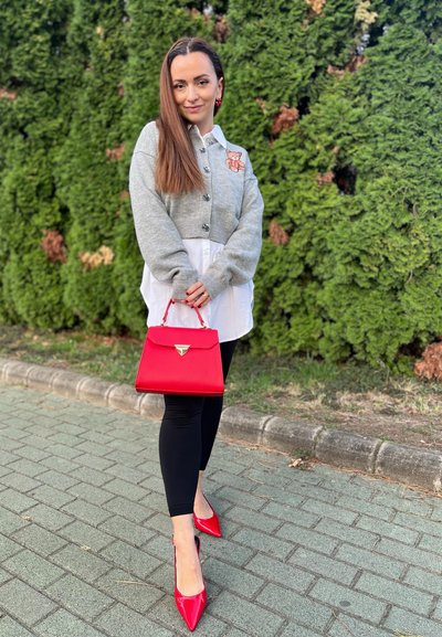 Cárdigan gris con parche de oso, camisa blanca, leggings negros y tacones rojos. Sosteniendo un bolso rojo con un acento triangular. Fondo verde.