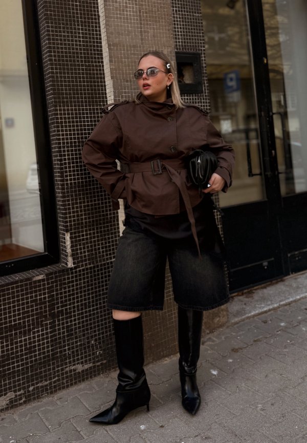 Frau trägt übergroße braune Jacke mit Gürtel, schwarze kniehohe Stiefel, Sonnenbrille und hält eine schwarze Clutch, steht auf einem gefliesten Bürgersteig.