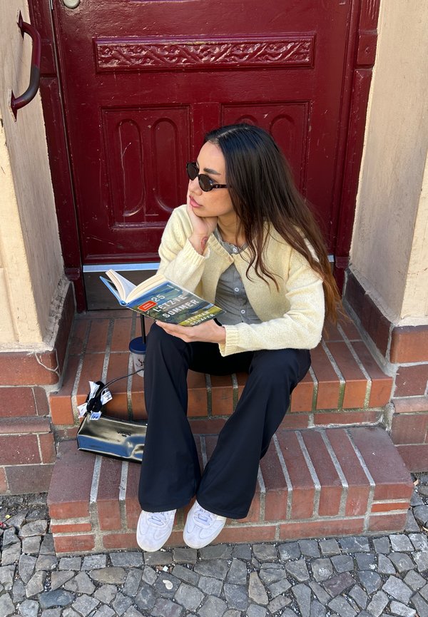 Jeune femme aux longs cheveux noirs et aux lunettes de soleil lit un livre assise sur des marches en briques devant une porte rouge, avec un sac noir à ses côtés.