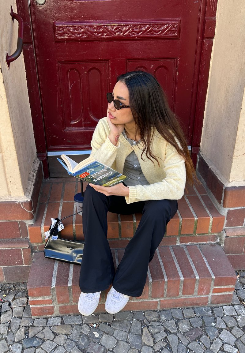 Jeune femme aux longs cheveux noirs et lunettes de soleil lit un livre assise sur des marches en briques devant une porte rouge, avec un sac noir à ses côtés.