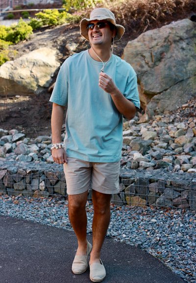 Hombre con gafas de sol, sombrero tipo pescador beige, camiseta azul claro y pantalones cortos caminando por un sendero con rocas y vegetación al fondo.