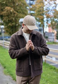 Brūns vilnas žakets ar divām priekšējām kabatām, pogu aizdari un augstu apkakli. Kombinēts ar bēša korduroja cepuri. Neitrālu toņu apģērbs.