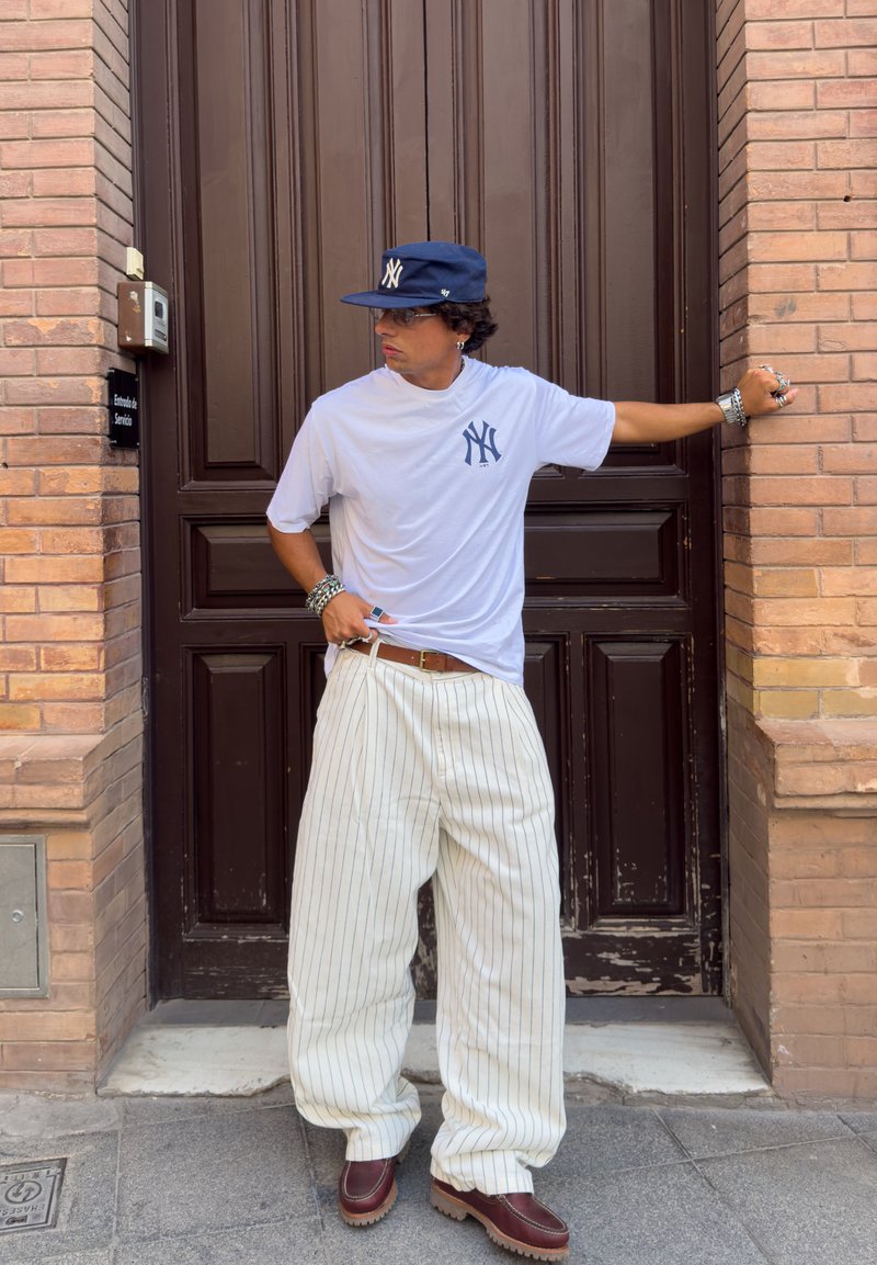T-shirt blanche avec un petit logo marine, pantalon large rayé beige et chaussures brunes à semelles épaisses. Les accessoires comprennent des bracelets et une casquette.
